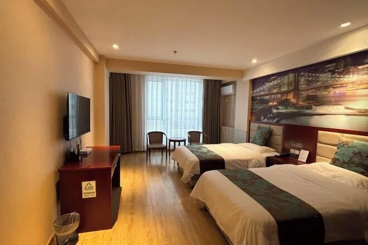 Quarto de hotel com duas camas, uma escrivaninha com televisão, uma cadeira, uma mesinha e uma janela grande com cortinas.