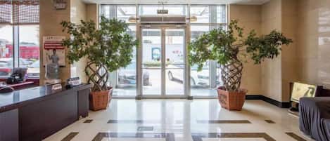 A hotel lobby with a reception desk, two potted plants, a glass door entrance, and a patterned floor.
