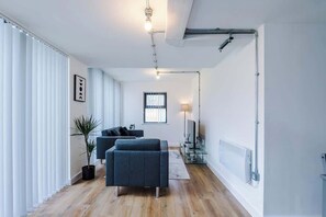 A modern living room with a sofa, a coffee table, and a TV. There is a window with blinds, a lamp, and a radiator. - Modern 2 Bedroom Apartment Near M6 Junction 21 (Warrington)