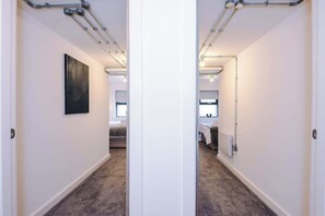 A hallway with a carpeted floor, a black artwork on the left wall, and a bed with a white headboard in the background. - Modern 2 Bedroom Apartment Near M6 Junction 21 (Warrington)