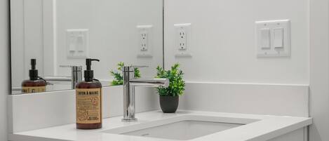 A modern bathroom with a white sink, a chrome faucet, a bottle of soap, and a potted plant.