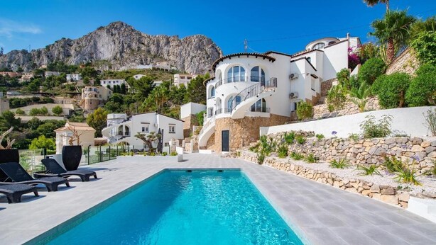 Une piscine entourée de chaises longues, un bâtiment blanc à la façade en pierre, et un paysage montagneux en arrière-plan.