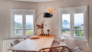 Un coin repas avec une table en bois, deux chaises, un vase de fleurs, une lampe fixée au mur et de grandes fenêtres offrant une vue sur un paysage montagneux.