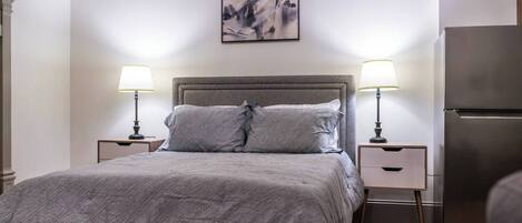 A bedroom with a bed, two bedside tables with lamps, a modern refrigerator, and a painting on the wall.