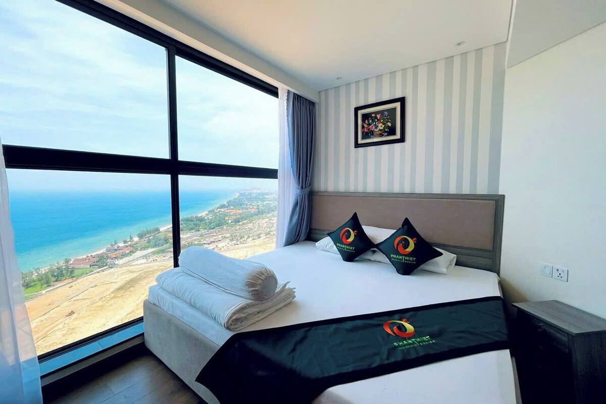 A hotel room with a large window offering a view of the sea and a distant landscape. The bed has a black bedspread with a logo, and there are two pillows with a similar logo.