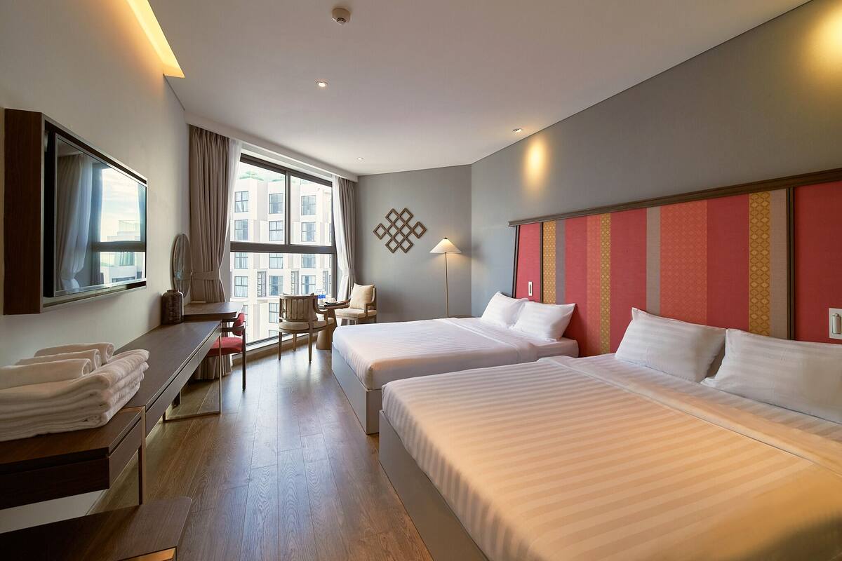 A hotel room with two beds, a desk with towels, a chair, a lamp, and a window with curtains.