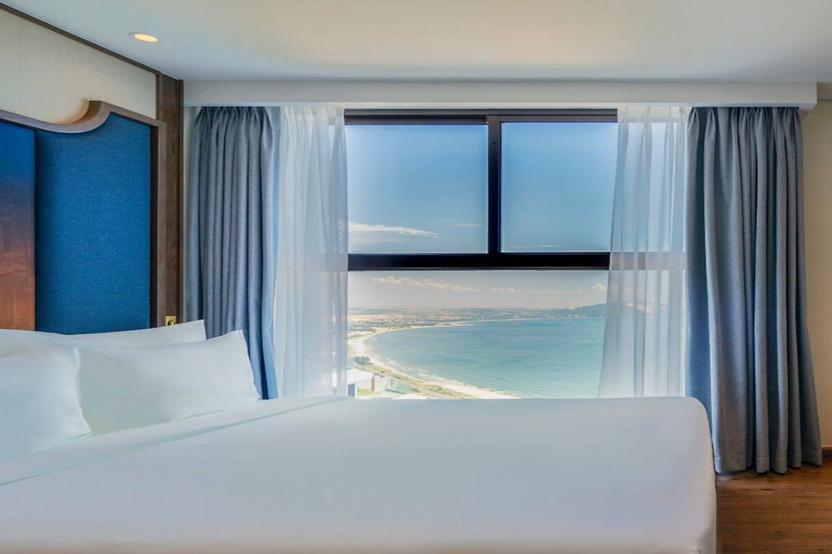 A hotel room with a large bed, a view of the sea, and a window with curtains.