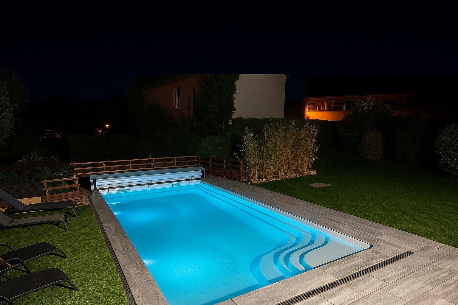 A rectangular swimming pool with a wooden deck and lounge chairs, surrounded by a well-maintained lawn and a hedge.