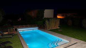 A rectangular swimming pool with a wooden deck and lounge chairs, surrounded by a well-maintained lawn and a hedge.