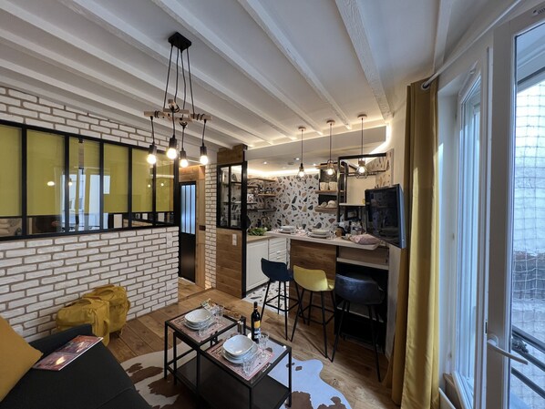 A modern living room with a sofa, coffee table, and dining area. There is a kitchen with a TV and a window with curtains.