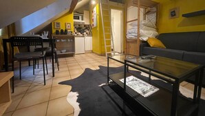 Een gezellige woonkamer met een glazen salontafel, een bank en een eethoek met stoelen, en een kleine keuken op de achtergrond.