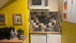 Een compacte keuken met een magnetron, spoelbak en een kleine koelkast. Er hangen afbeeldingen in lijsten aan de muur en is een betegelde spatwand.