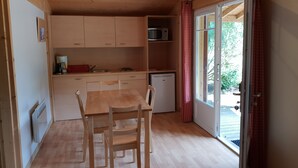Cocina compacta con armarios de madera, microondas, pileta y heladera pequeña. Hay una mesa de comedor con sillas y una ventana con cortinas.