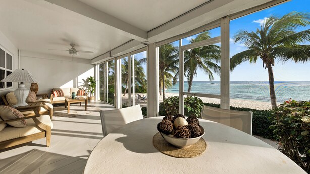 Een ruime kamer met een groot raam met uitzicht op een strand, ingericht met een bank, stoelen en een eettafel met een schaal decoratieve objecten.