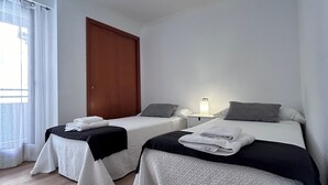 A hotel room with two beds, a wooden floor, a window with white curtains, and a door.