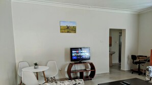 Een woonkamer met een flatscreen televisie, witte stoelen, een bloemenbank en een donkere salontafel.