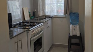 Een keuken met witte kastjes, een gasfornuis, een oven, een raam met gordijn en een waterdispenser.