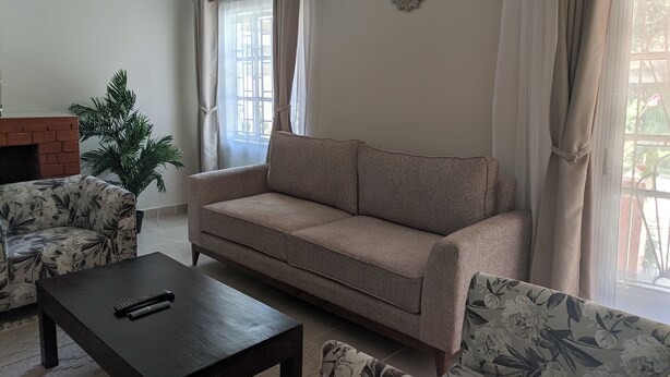 Een woonkamer met een bank, twee fauteuils, een salontafel, een open haard en een groot raam met gordijnen.