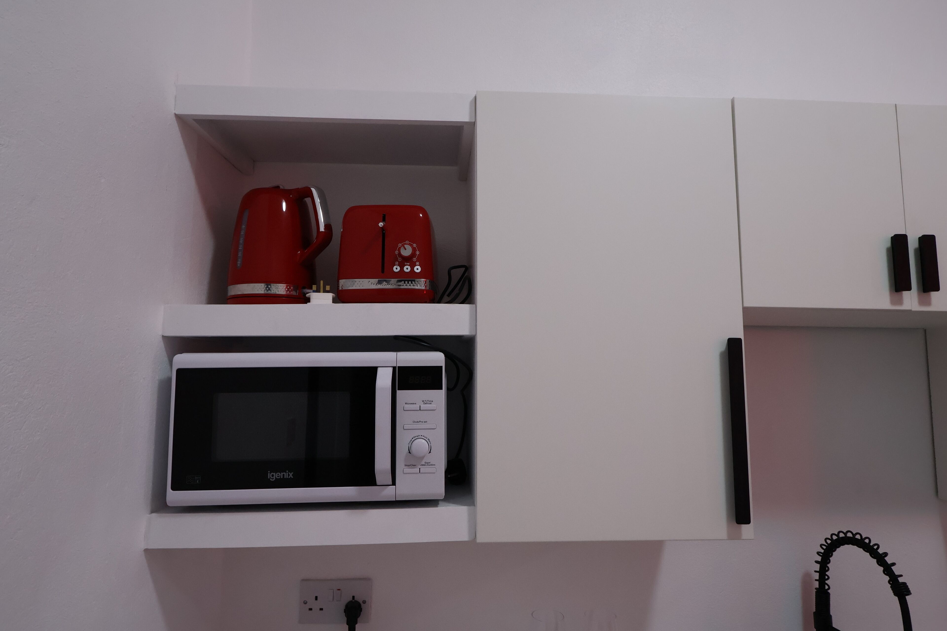 Una cocina con un microondas blanco, una cafetera roja y un armario blanco.