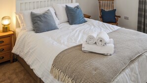 Un lit bien fait, avec du linge de lit blanc, un plaid beige et des serviettes pliées. On y trouve une table de chevet en bois avec une lampe, une chaise avec un coussin bleu et une tête de lit blanche.