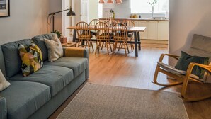 Un salon moderne avec un canapé gris, une table à manger en bois et des chaises, un tapis à motifs chevrons et une cuisine en arrière-plan.