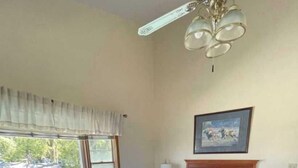A hotel room with a bed, a table, a chair, a ceiling fan, a window with curtains, and a painting on the wall.