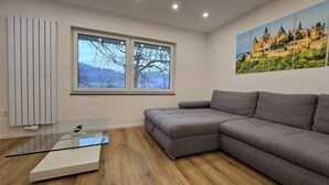 Een moderne woonkamer met een grijze bank, een glazen salontafel en een groot raam met uitzicht op bomen en heuvels.