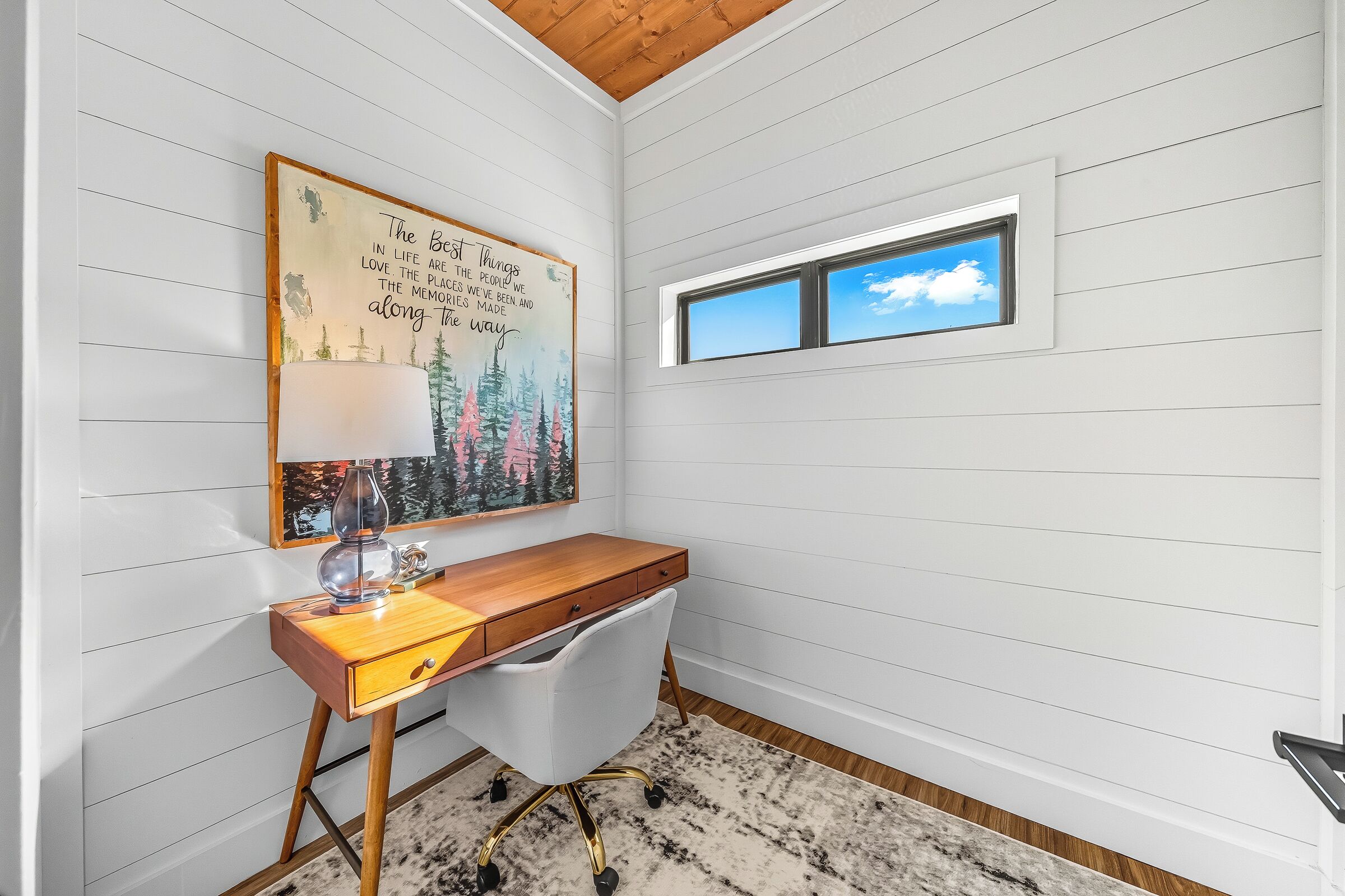 A room with a desk, a lamp, a painting, a window, and a ceiling with wooden beams.