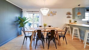 A dining area with a wooden table, black chairs, a chandelier, and a kitchen area with stools. - Cozy Riverside Cottage—Quiet—3-Beds—Fully Updated! (Belmont)