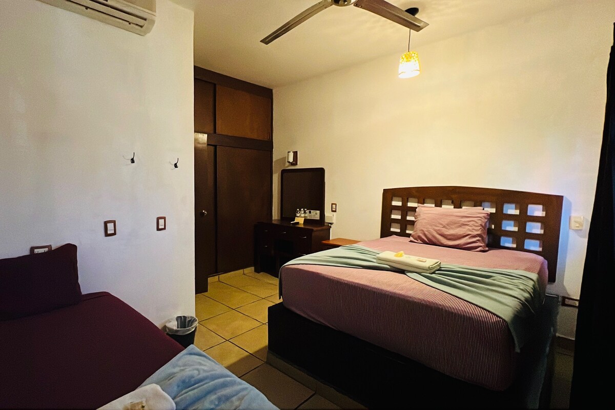 A hotel room with two beds, a wardrobe, a mirror, a ceiling fan, and a small table with a lamp.