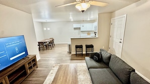 A modern living room with a flat-screen TV, a gray sofa, and a wooden coffee table. There is a dining area with a table and chairs, and a kitchen in the background.