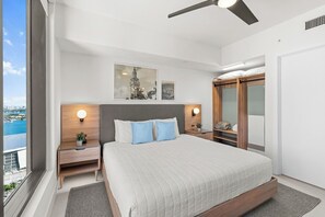 A modern bedroom with a large bed, two bedside tables with lamps, a ceiling fan, and a view of the city through a large window.