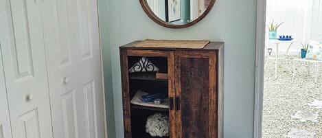 Una habitación con una puerta blanca, un mueble de madera con estantes, un espejo redondo y un cubo de basura azul.