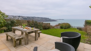 A patio with a wooden table set for dining, wicker chairs, and a view of the sea. - Heatherleigh, Carbis Bay – Sleeps 8 – Sea Views – Dog-Friendly - On-Site Parking (Carbis Bay)