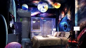 A bedroom with a bed, a desk, a chair, and a view of the cityscape through a window. - Aegean Sea Theme Hotel (Nanping)