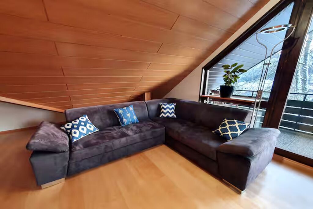 Un salon moderne avec un canapé d’angle marron foncé, un plafond en bois à pente et une grande fenêtre donnant sur un paysage enneigé.