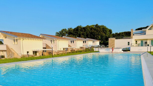 Une piscine dotée d’une terrasse carrelée, entourée de bâtiments modernes à la façade en pierre, sous un ciel d’un bleu éclatant.