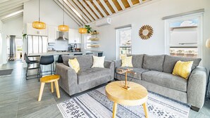 En rummelig opholdsstue med en grå sofa, et rundt træbord og et køkken i baggrunden.