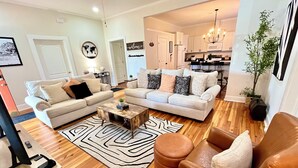 A modern living room with a sofa, coffee table, and ottoman, wooden flooring, and a kitchen in the background. - NEW-Heart of Kannapolis-Modern & Charming Escape (Kannapolis)