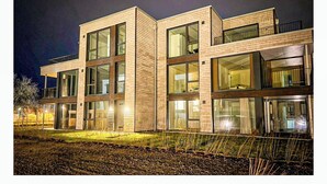 A modern multi-story building with large windows and a flat roof, featuring a brick facade and a well-lit entrance. - Apartment BeltEck - sea view. Sauna. Family-friendly. Dogs on request (Fehmarn)