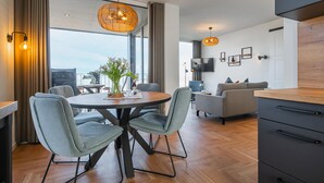 A modern living room with a round dining table, grey upholstered chairs, a grey sofa, a TV mounted on the wall, and a large window with curtains. - Apartment BeltEck - sea view. Sauna. Family-friendly. Dogs on request (Fehmarn)