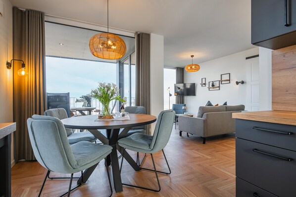 A modern living room with a round dining table, grey upholstered chairs, a grey sofa, a TV mounted on the wall, and a large window with curtains.