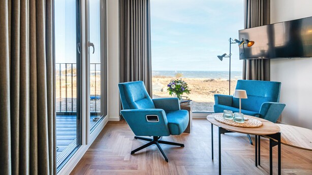 A modern hotel room with a large balcony, a flat-screen TV, and a seating area with two blue chairs and a small table. - Apartment BeltEck - sea view. Sauna. Family-friendly. Dogs on request (Fehmarn)