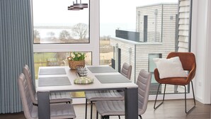 Ein moderner Essbereich mit einem Tisch für vier Personen, einem braunen Stuhl mit weißem Kissen und Blick auf ein Haus durch ein großes Fenster.