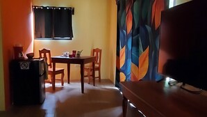 Una habitación con un banco de madera, una mesa con sillas, un refrigerador y un ventanal con cortinas.