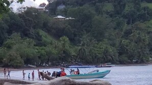 Una playa con un bote amarrado cerca de la orilla, rodeada de abundante vegetación y algunas personas.