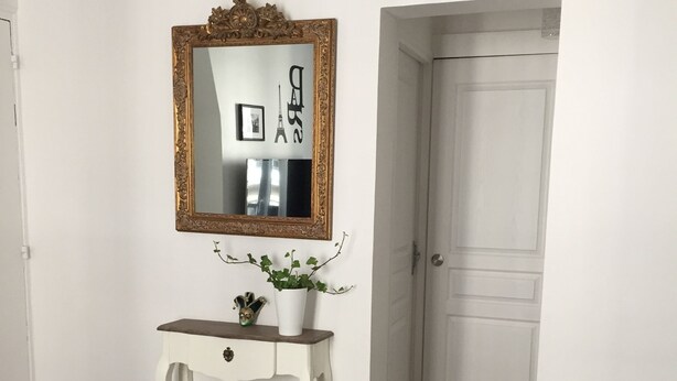 Ein Zimmer mit weißem Konsolentisch, einem Spiegel mit goldener Rahmung, einer Topfpflanze und einem grauen Sofa.