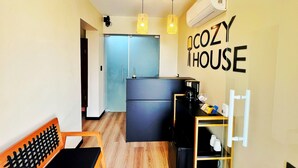Una cocina compacta con refrigerador, cafetera y repisa con tazas. Se ve un banco y una silla, y la habitación tiene un letrero en la pared que dice 'COZY HOUSE'.