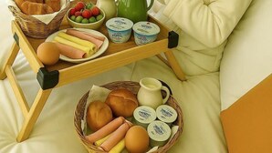 Una persona sentada en una cama con una bandeja de alimentos para el desayuno, incluyendo pan, huevos y salchichas.