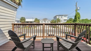 Una terraza de madera con dos sillas Adirondack y una mesa pequeña, con vista a una calle con casas y un cielo despejado.